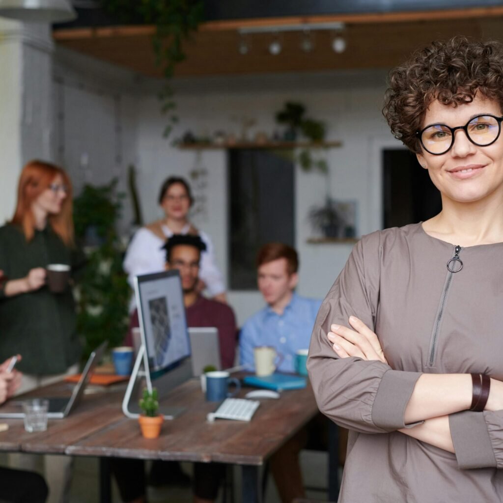 Smiling businesswoman with curly hair stands confidently in a modern office space with colleagues.